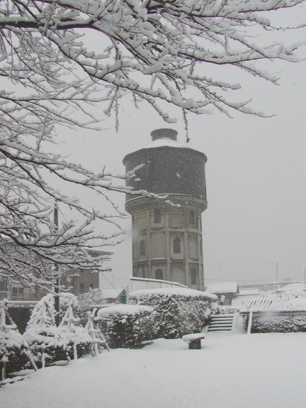 雪が降っている風景の中に雪の積もった屋根が複数ある中下が灰色で上が茶色の2色の塔のような建物の写真