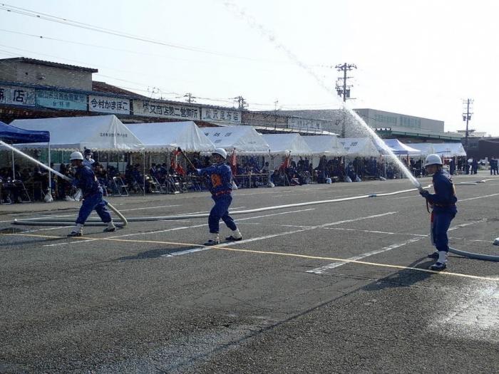 奥にテントが幾つか立ち並んでおり、その手前では3人のヘルメットをかぶった団員らがホースを使って放水を行っている様子の写真