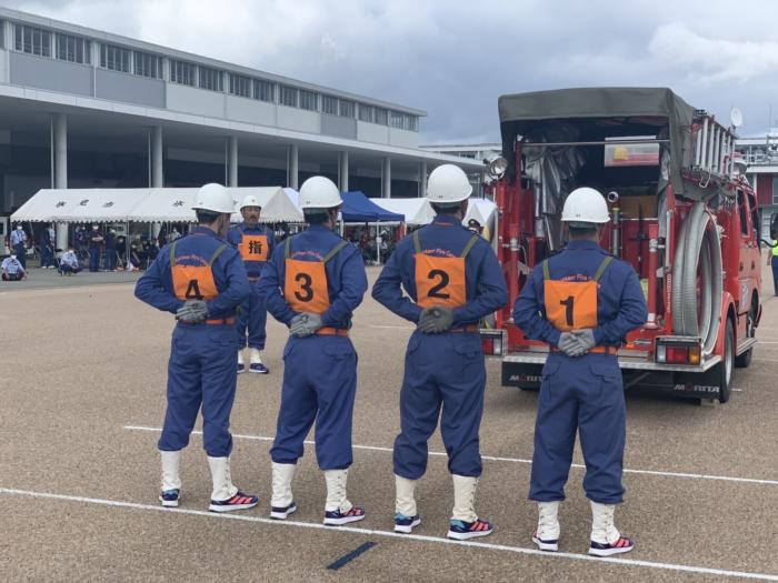 手前に、右から1・2・3・4と数字のゼッケンとヘルメットを身に纏った4人の団員が後ろで手を組んで並び、その奥に「指」と書かれたゼッケンとヘルメットを身に纏った団員が立っており、その右隣に消防車が止まっている様子の写真