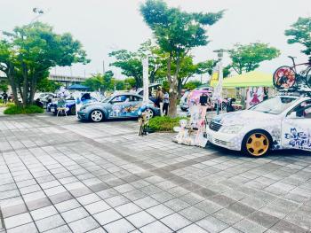 手前に、白地に青い花をあしらった塗装で、屋根に自転車を載せた車があり、その向こうに青地にイラストの描かれた車が見える写真