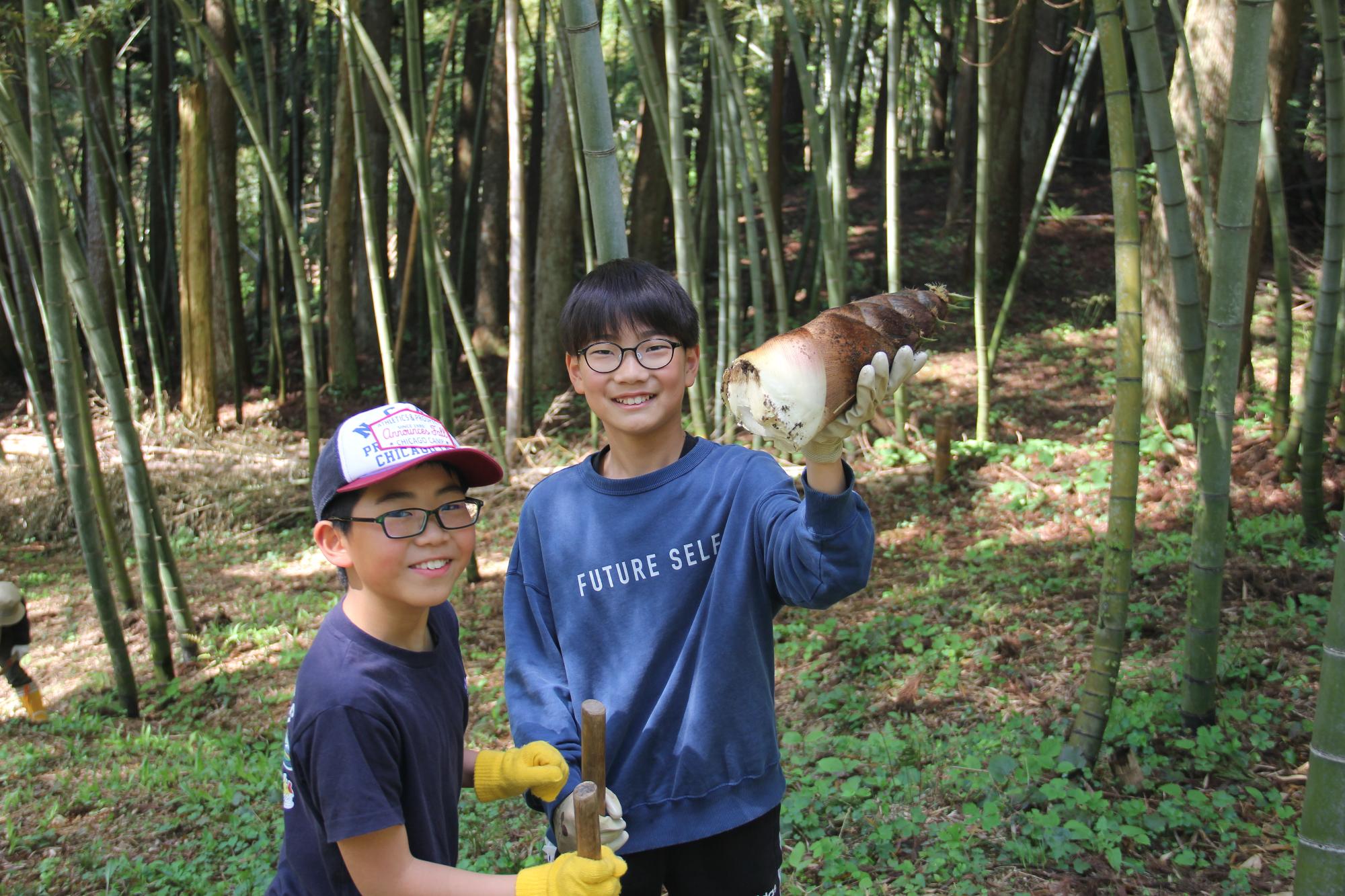 笑顔で採れたての大きな竹の子を掲げる兄弟の様子