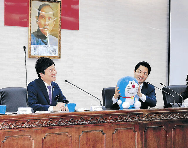どらえもんのぬいぐるみを両手でつかみにこやかに話す中国台北市のショウ市長とその隣で笑顔をみせる角田市長